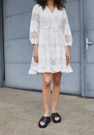 Persona che indossa un vestito bianco in pizzo sangallo con maniche a sbuffo e sandali neri con plateau, in piedi su un pavimento di cemento vicino a una porta grigia.