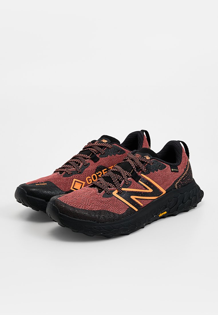Par de zapatillas de trail running New Balance en rojo y negro con detalles naranjas, tejido Gore-Tex y suelas Vibram sobre fondo blanco.