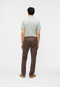 Άνδρας που στέκεται με την πλάτη, φοράει ανοιχτό γκρι polo με κοντά μανίκια, καφέ παντελόνι και ταμπά παπούτσια, μπροστά σε λευκό φόντο.