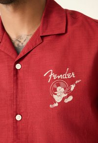Chemise rouge à boutons avec logo Fender blanc et une illustration d'un personnage de disque vinyle jouant de la guitare électrique sur la poitrine.