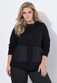 Pull noir à col volants, manches longues et deux grandes poches en tissu. Un pantalon rayé noir et blanc complète la tenue.
