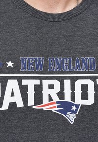 Camiseta de algodón gris con la inscripción "NEW ENGLAND PATRIOTS" en texto blanco y azul, con un logotipo gráfico de la cabeza de un patriota en rojo, blanco y azul.