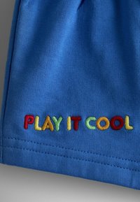 Blåt stof med teksten "PLAY IT COOL" broderet i flerfarvede bogstaver nær den syede kant.