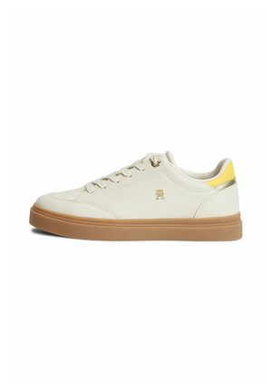 Witte lage sneaker met rubberen zool, witte veters, klein gouden logo aan de zijkant en een geel accent op de hiel.