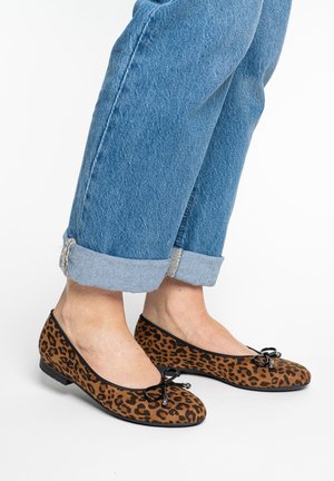 Benen in opgerolde blauwe spijkerbroeken, dragen bruine ballerina's met luipaardprint, zwarte rand en kleine strikjes, op een witte achtergrond.