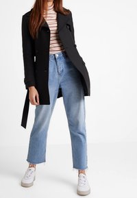 Žena v černém dvouřadém kabátu, pruhovaném roláku, světle modrých džínech s roztrhanými okraji a bílých teniskách stojíc�í na bílém pozadí.