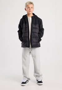 Musta pehmeä takki, jossa on vetoketju edessä, huppu ja kaksi etutaskua. Puettu kevyen paidan ja harmaiden verkkareiden päälle. Rentouttavat lenkkarit.