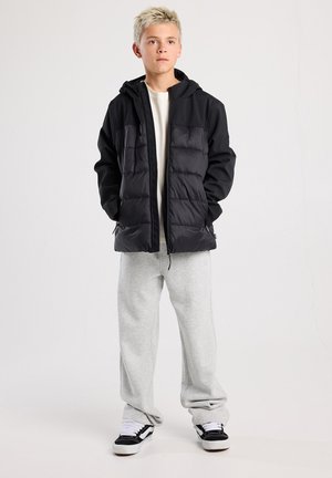 Schwarze gepolsterte Jacke mit Reißverschluss, Kapuze und zwei vorderen Taschen. Getragen über einem leichten Hemd und grauen Jogginghosen. Legere Sneaker.