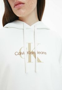 Felpa bianca con cappuccio, con cordino, che presenta un logo ricamato dorato "CK" e "Calvin Klein Jeans" sottostante in un font a contrasto.
