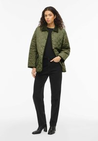Veste matelassée vert olive avec col, fermeture à bouton sur le devant et poches latérales, associée à un jean noir droit et des bottines noires.