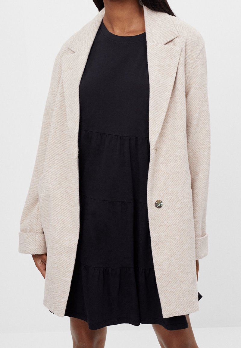 Femme portant un blazer beige oversize sur une robe noire à étages longueur genou, debout devant un fond clair uni.