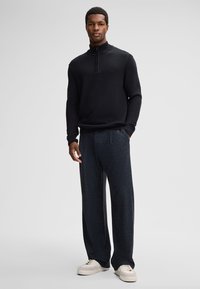 Zwarte gemêleerde zip-nek trui gecombineerd met donkere, gemusterde sweatpants en beige sneakers. Model staat met handen in de zakken.