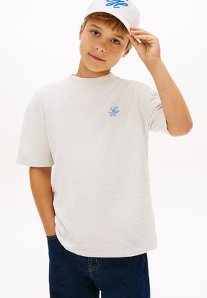 Dreng iført en hvid tekstureret kortærmet skjorte og matchende hvid kasket med blåt logo, poserer med den ene hånd, der justerer kasketten.