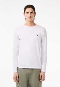 Hvid langærmet t-shirt lavet af blød bomuld, med et lille grønt logo på brystet. Enkel rund hals, glat tekstur og tætsiddende design.