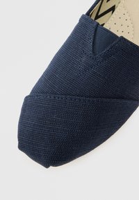 Granatowy, płócienny but wsuwany o fakturowanym wykończeniu, z trójkątnym przeszyciem jako akcentem oraz beżową, perforowaną wnętrzem.