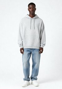 Pelēka kokvilnas kapuce ar priekšējo kabatu, regulējamu kapuci un ribotām piedurknēm, apvienota ar gaiši ziliem džinsiem un baltām sporta apaviem.
