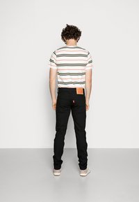 Camisa de manga corta a rayas en blanco, verde y rosa; combinada con vaqueros negros que llevan un parche con logo. Zapatos lisos y claros completan el conjunto.