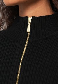 Μαύρο ριμπ πουλόβερ με ψηλό λαιμό και λεπτομέρεια από χρυστό φερμουάρ στο μπροστινό μέρος. Το ύφασμα έχει υφή που αναδεικνύει το πλεκτό μοτίβο του.