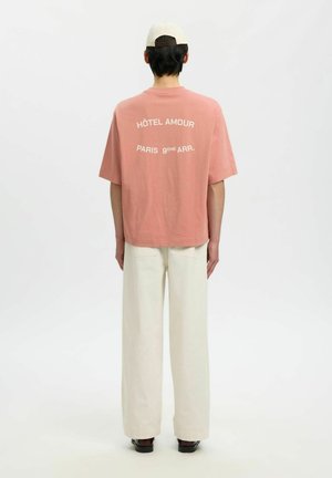 Isik, kes kannab seljas lõhesinine T-särki, millel on taga kiri "HÔTEL AMOUR PARIS 9ÈME ARR.", kreemikaid laiade säärega pükse, musti jalatseid ja beeži lippmütsi, seisab seljaga vaataja poole.