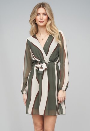 Ung kvinde med langt blondt hår iført en grøn, beige og brun stribet slå-om kjole med lange gennemsigtige ærmer og et stofbælte.