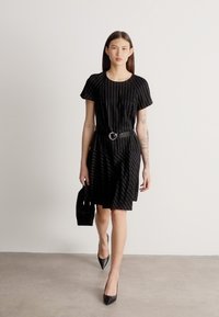 Moteris vilki juodą dryžuotą suknelę su diržu, laiko juodą rankinę ir juodus aštriais galiukais aukštakulnius, stovėdama priešais paprastą foną.