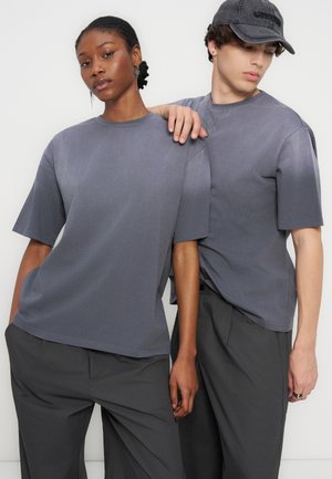 Δύο μοντέλα με ταιριαστά γκρι oversized t-shirts και σκουρόχρωμο παντελόνι, ένα με καπέλο, ποζάρουν σε απλό φωτεινό φόντο.