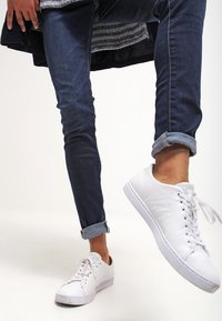 Zapatillas deportivas blancas con tejido texturizado, puntera redonda y cordones; combinadas con jeans oscuros de tiro alto y remangados. Diseño simple y casual.