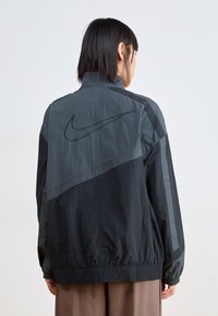 Černá a tmavě šedá větruodolná bunda s vysokým límcem, elastickými manžetami a lemem. Na zadní straně se nachází nenápadné logo Nike. Lehký materiál.