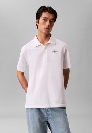 Rövid fekete hajú fiatal férfi fehér Calvin Klein galléros ingben és világoskék farmerral, egyszínű szürke háttér előtt áll.