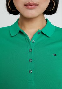 Zielona koszulka polo z teksturowanej tkaniny z kołnierzykiem, z trzema czarnymi guzikami i małym haftowanym logo po lewej stronie.
