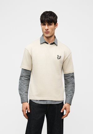 Άνδρας με μαύρα μαλλιά που φοράει μπεζ κοντομάνικο πουκάμισο πάνω από μακρυμάνικο πουκάμισο ασπρόμαυρη καρό, μαύρο παντελόνι, στέκεται μπροστά σε λευκό φόντο.