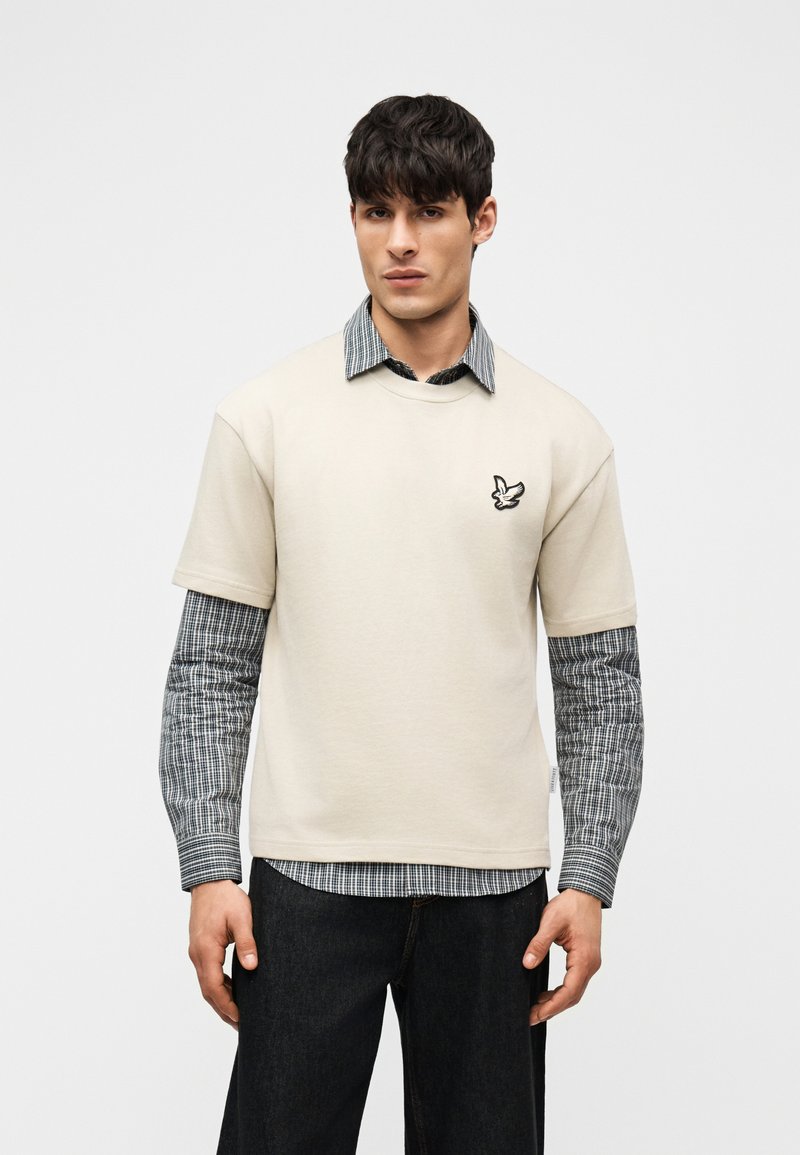 Άνδρας με μαύρα μαλλιά που φοράει μπεζ κοντομάνικο πουκάμισο πάνω από μακρυμάνικο πουκάμισο ασπρόμαυρη καρό, μαύρο παντελόνι, στέκεται μπροστά σε λευκό φόντο.