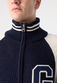 Námornícky modrý zip-up sveter s vysokým rebrovaným golierom, bielymi pruhmi a textúrovaným pletením. Na prednej strane je veľké, svetlé písmeno "G".