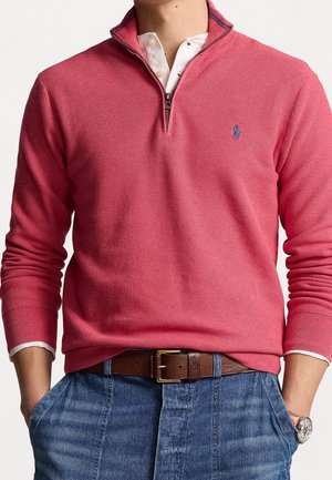 Homme portant un pull rose à fermeture éclair quart, sur une chemise blanche à col, jean bleu, ceinture marron et montre-bracelet, mains dans les poches.