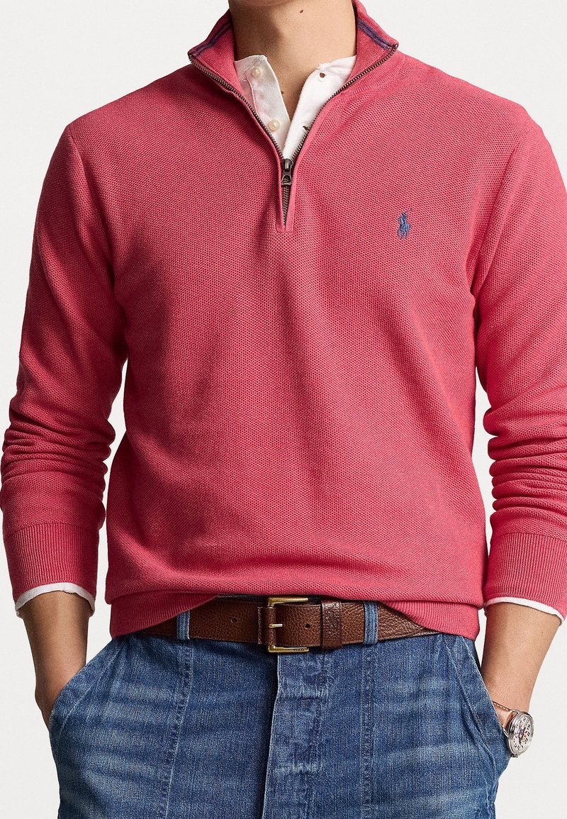 Homme portant un pull rose à fermeture éclair quart, sur une chemise blanche à col, jean bleu, ceinture marron et montre-bracelet, mains dans les poches.