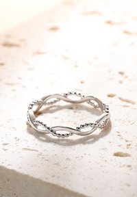 Srebrny zakręcony pierścionek z teksturowanymi obręczami i perłowymi akcentami, wyeksponowany na jasnym kamiennym podłożu.