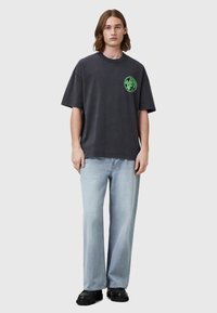 Løst siddende charcoal t-shirt med rund halsudskæring og grøn grafisk logo på brystet, parret med lyseblå jeans og sort fodtøj.