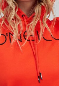 Gros plan d'une personne aux cheveux blonds longs portant un sweat à capuche orange vif avec un logo O'Neill noir et des cordons de serrage.
