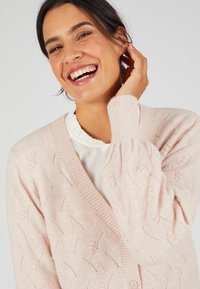 Cardigan in rosa chiaro lavorato a maglia con scollatura a V, motivo a trama e polsini a coste, abbinato a una camicia bianca con volant sotto.