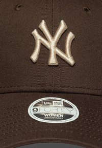 Gorra ajustable marrón con un logo bordado en beige de "NY", que presenta una visera curva y un tejido texturizado. Incluye una etiqueta redonda con los detalles de la marca.