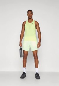 Atletski muškarac stoji, nosi neonsko žutu majicu bez rukava i kratke hlače, bijele Nike čarape, crne tenisice, drži crnu bocu vode.