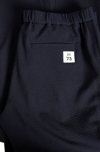 Temno modri atletski šortai iz teksturirane mrežaste tkanine. Imajo elastičen pas in zadnji žep z belim etiketom, na katerem je napisano "M 73."