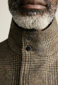 Pruun ruuduline villane mantel kõrge krae ja kahe silmapaistva metalsinise nupuga, millel on tekstureeritud viimistlus. Üksikasjalik vaade krae detailile.