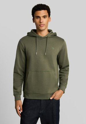 Junger Mann trägt einen olivgrünen Kapuzenpullover mit Fronttasche und dunkle Jeans, stehend vor einem einfarbigen grauen Hintergrund.