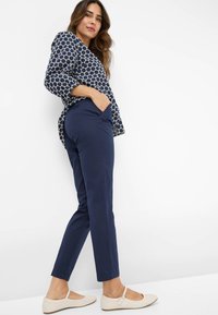 Bluse mit maritimen Mustern und kreisförmigen Designs, kombiniert mit marineblauen Slim-Fit-Hosen und hellbeigen flachen Schuhen, die ein lässiges Outfit zeigen.