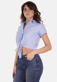 Camicia azzurra a maniche corte con righe bianche, annodata in vita. Presenta un colletto a punta e chiusura con bottoni. Abbinata a jeans di denim a vita alta.