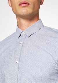 Světle modrá košile na knoflíky s texturovaným vzorem, která má klasický límeček a bílé knoflíky. Detailní pohled na horní část oděvu.