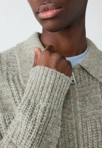 Zbliżenie na dolną część twarzy oraz rękę osoby, która poprawia kołnierz beżowego, strukturalnego swetra z zamkiem błyskawicznym.