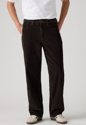 Homme portant un pantalon en velours côtelé noir et des baskets blanches à rayures vertes sur un fond clair uni.