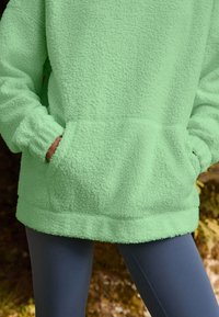 Green fleece pullover with a textured surface, two front pockets, and a loose fit. Paired with dark leggings, showing a layered look.